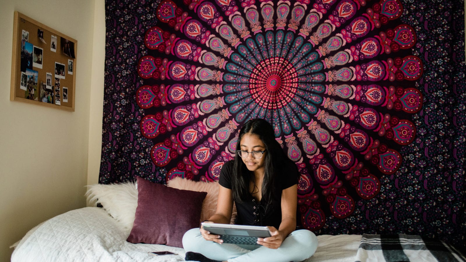 girl with glasses in dorm room sitting on bed in front of wall hanging