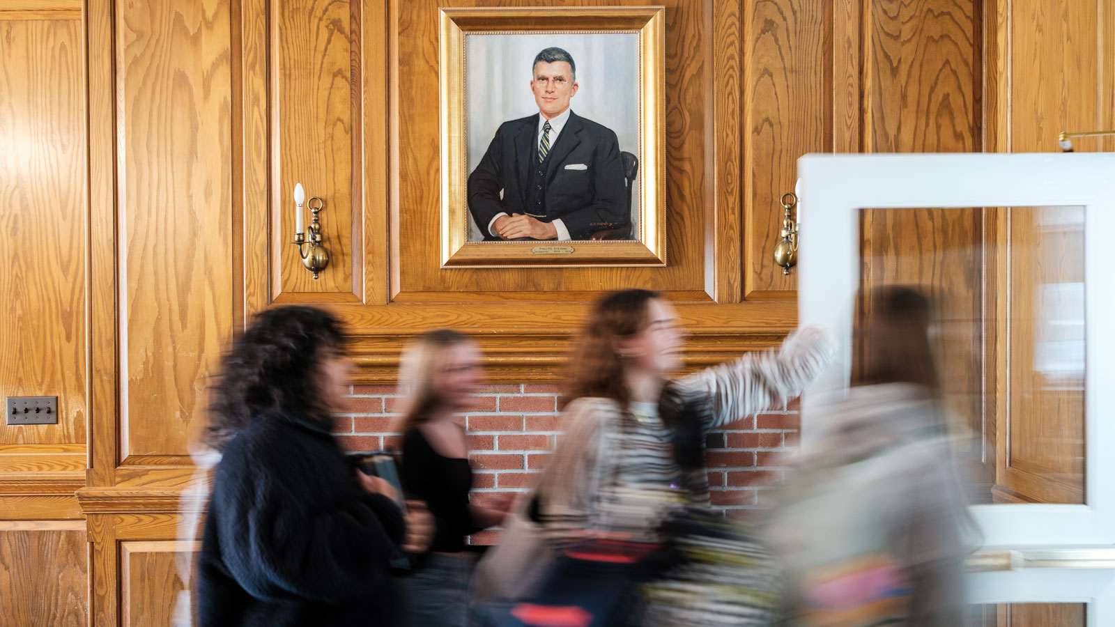Students walking by portrait in wood-paneled hallway