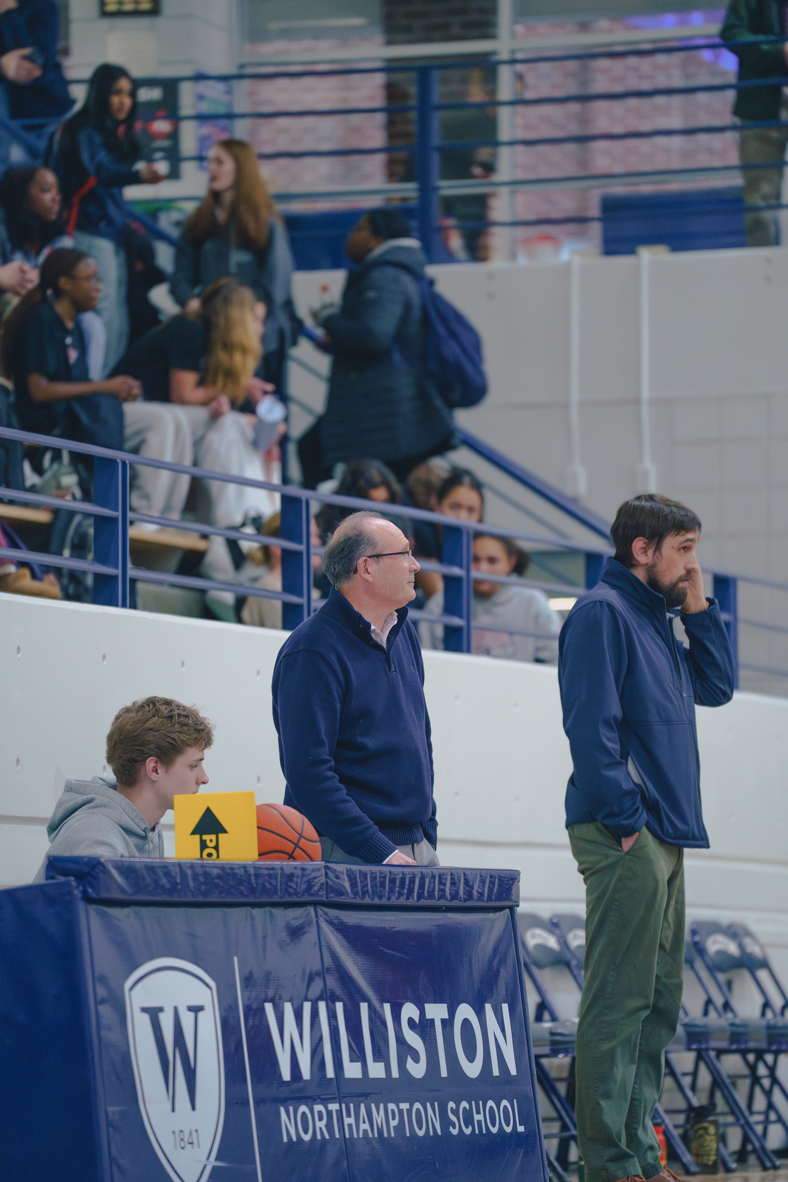 Two Williston coaches stand near the basketball scorers table, one Williston student sits