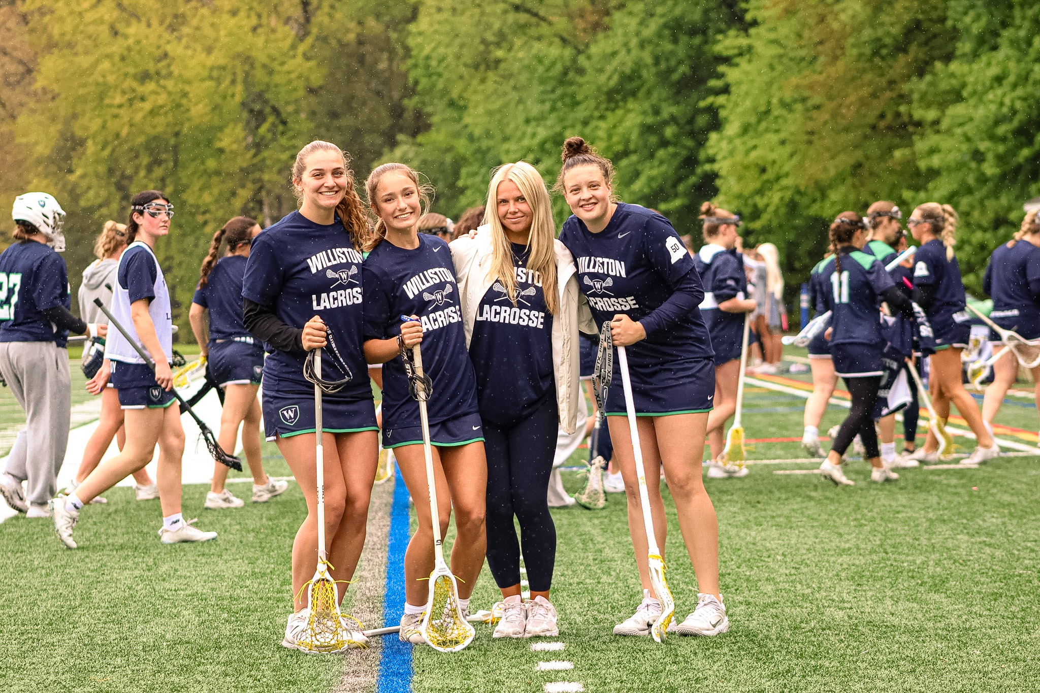 Four girls lacrosse players pose for picture on sideline during a game