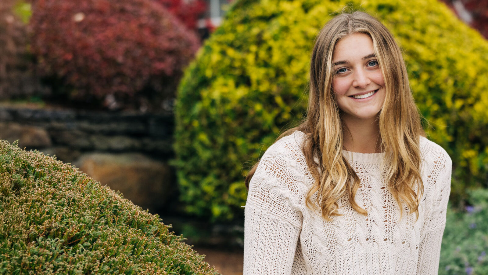 Female student in sweater sitting in front of bushes